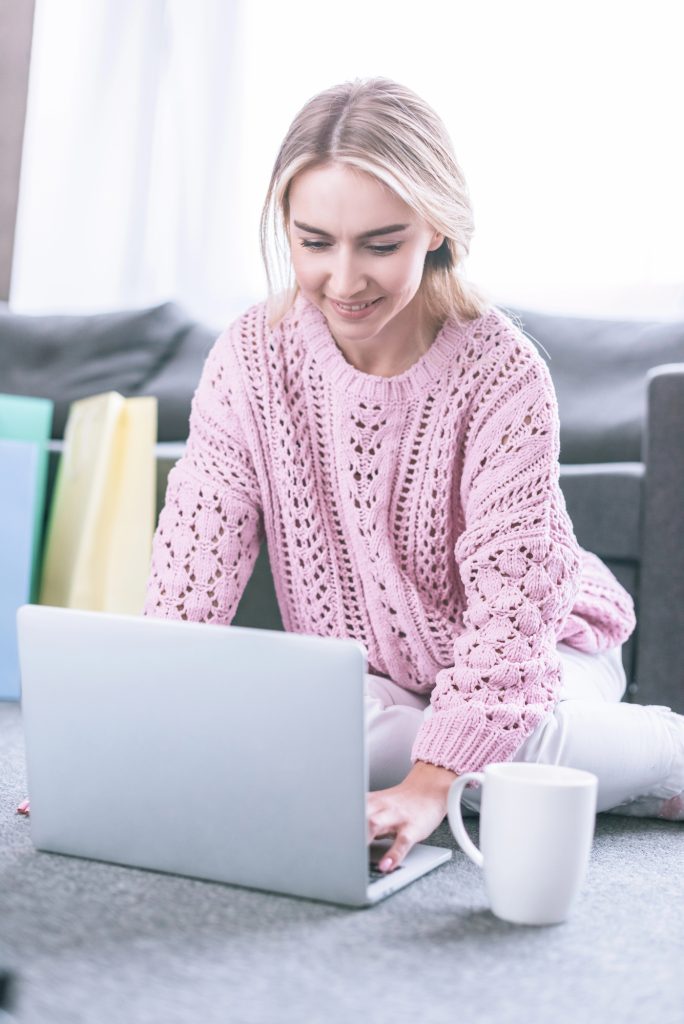 A person in a pink sweater sits on the floor with a laptop, sipping from a white mug. They're planning to turn their blog into a book, capturing their thoughts in every keystroke.
