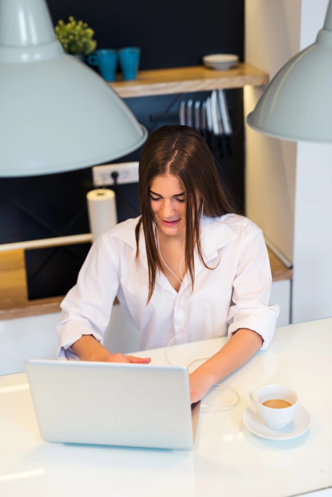 A woman in a white shirt sits at a table, using her laptop to start a career as a ghostwriter. A cup of coffee sits nearby, fueling her creativity, while kitchen items subtly frame the background.