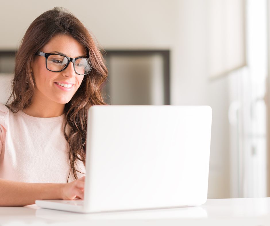 Woman with brown hair and wearing glasses working on a laptop while smiling for the blog post "the importance of social media for freelance ghostwriters"